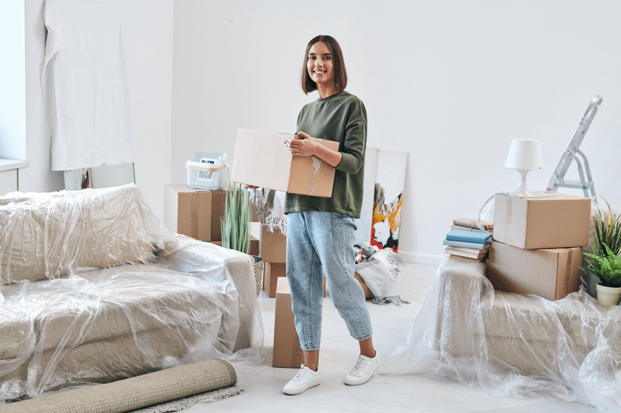 Move In Smoothly At The Johnny - Cheerful young woman in jeans and a sweatshirt carrying a packed box while moving into a new flat or house in front of the camera.